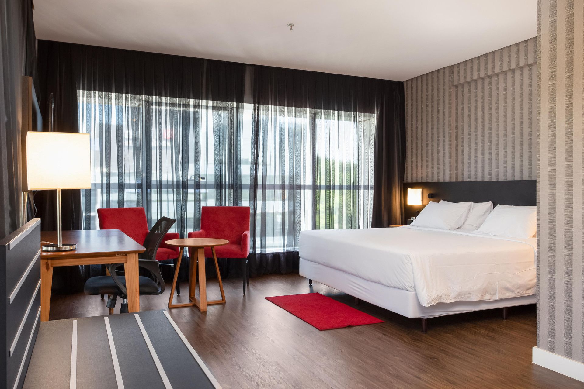 Hotellihuone, jossa on sänky, työpöytä lampun kanssa, kaksi tuolia ja näkymä suurista ikkunoista.