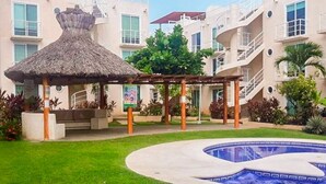 Une piscine agrémentée d’un kiosque à toit de chaume, entourée d’une pelouse bien entretenue et d’un immeuble à plusieurs étages doté de balcons.