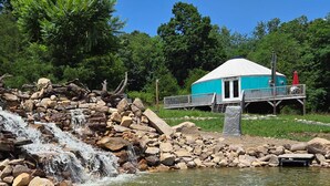 A turquoise yurt-style structure with a white roof, situated on a wooden deck over a rocky area with a waterfall in the foreground. - Relax and unwind at Solar Solitude (Mathias)