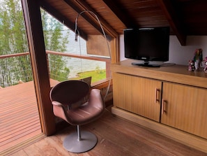 A modern room with a wooden ceiling, a television on a wooden cabinet, a chair, and a view of a lake through large windows. - Cabana à Beira D'água Linda Vista (Imbituba)