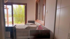 A bedroom with a large bathtub, a bed with striped bedding, a window with a view of greenery, and a wooden door. - Cabana à Beira D'água Linda Vista (Imbituba)