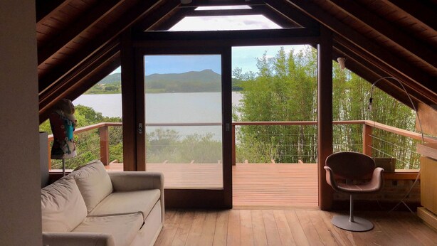 An attic room with a wooden floor, a sofa, a chair, and a large window overlooking a lake and greenery. - Cabana à Beira D'água Linda Vista (Imbituba)