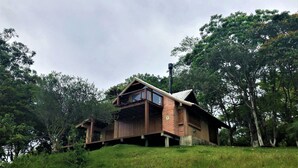 A wooden cabin with a balcony, surrounded by trees and grassy area. - Cabana à Beira D'água Linda Vista (Imbituba)