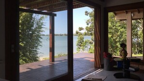 A room with a large sliding glass door leading to a deck with a view of a lake and trees. - Cabana à Beira D'água Linda Vista (Imbituba)