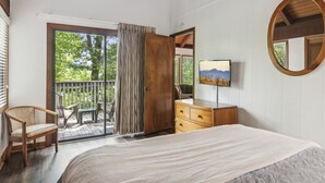 A bedroom with a large bed, a wooden dresser, a chair, a television, and a balcony with a view of trees. - Petit Crest Villas (Jasper)