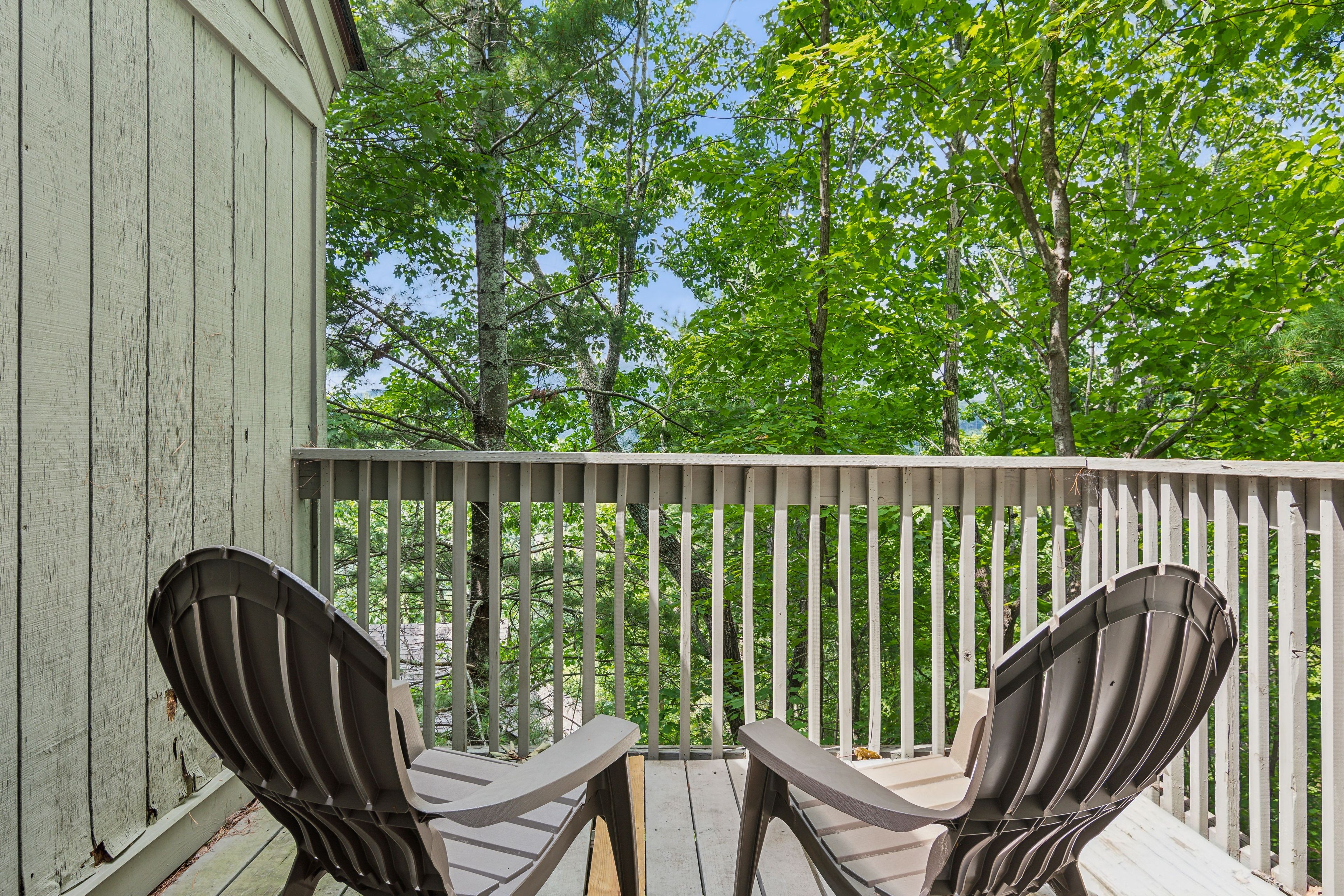 Une terrasse en bois avec deux chaises et une table, entourée d’une balustrade blanche, donnant sur une zone boisée.