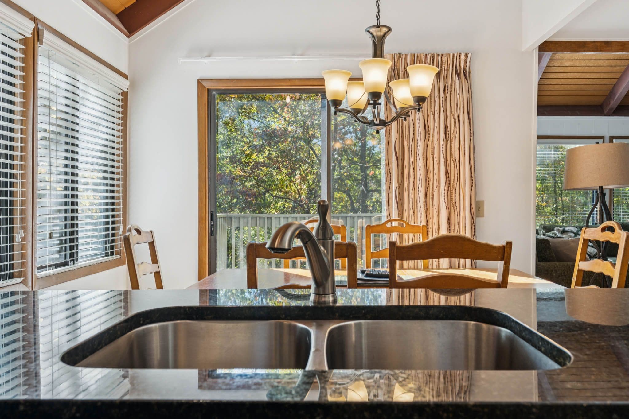 A kitchen with a double sink, wooden chairs, a chandelier, and a view of trees through large windows.