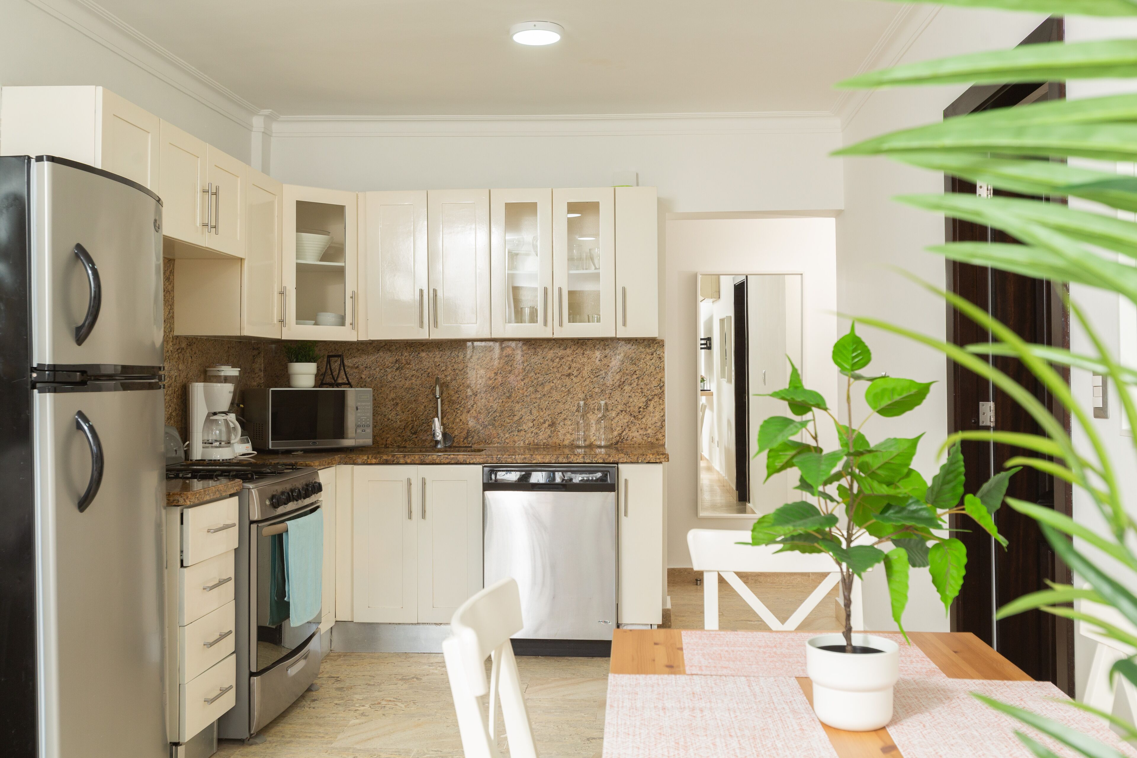 Une cuisine compacte avec des meubles blancs, un réfrigérateur en acier inoxydable, un four à micro-ondes, un four et un lave-vaisselle. On y trouve une table en bois sur laquelle est posée une plante en pot.