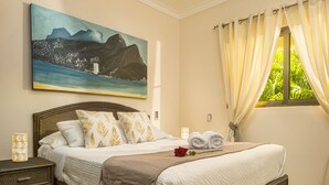 A neatly made bed with a white quilt, a brown throw blanket, and a red rose. There is a painting of a coastal scene on the wall, and a window with curtains. - Playa Laguna Beach Condos (Cabarete)