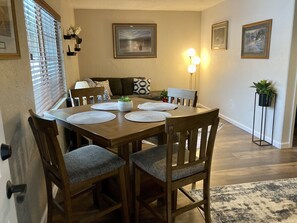 Un comedor con una mesa de madera, cuatro sillas y una alfombra. Hay cuadros en las paredes y una planta sobre un soporte.