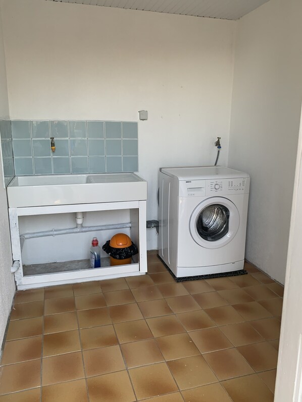 Une petite buanderie comprenant un lave-linge, un évier avec un pare-éclaboussures carrelé et une poubelle.