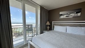 Hotelkamer met een groot bed, nachtkastje, lamp en uitzicht op de oceaan door een schuifdeur.
