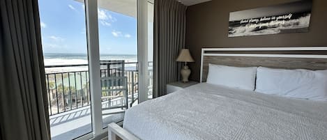 A hotel room with a large bed, a nightstand, a lamp, and a view of the ocean through a sliding glass door.