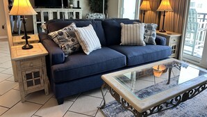 Een woonkamer met een donkerblauwe bank, twee bijzettafels met lampen, een glazen salontafel en een televisie op een meubel.