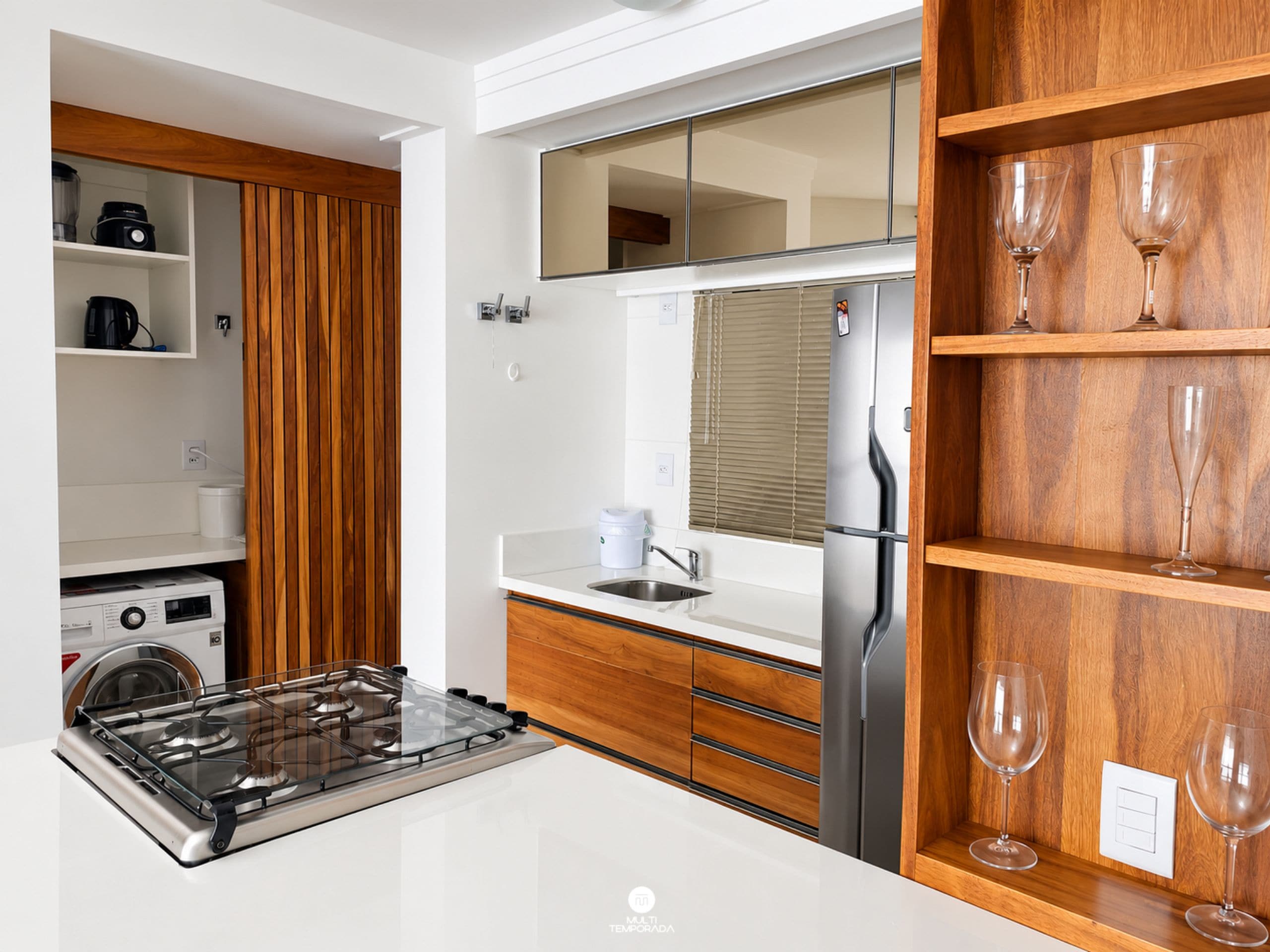 Una cocina moderna con estufa integrada, refrigerador, estantes de madera con copas de vino y lavadora al fondo.