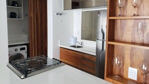 A modern kitchen with a built-in stove, a refrigerator, and wooden shelves with wine glasses. - Apartamento de Frente pro Mar - Campeche (Florianópolis)