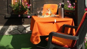 An outdoor dining area with a table set for two, a chair, and a potted plant with pink flowers. - Alpha Hotel (Saas Grund)