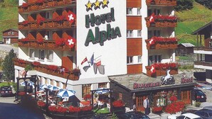 A multi-story hotel with balconies adorned with red flowers and Swiss flags, a restaurant, and a parking area. - Alpha Hotel (Saas Grund)
