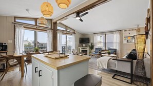 Un salon moderne avec un canapé, un coin repas comprenant une table et des chaises, une cuisine avec un comptoir et de grandes fenêtres offrant une vue sur l’extérieur.