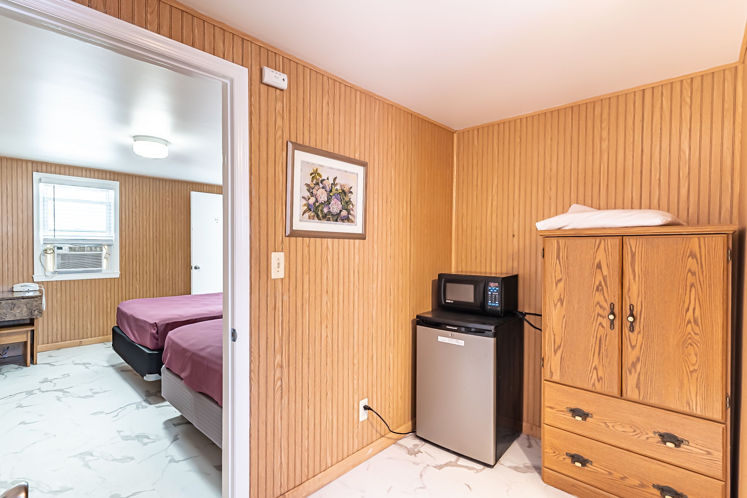Una habitación compacta con paneles de madera, una cama con ropa de cama rosa, un refrigerador pequeño, un microondas y un mueble con cajones.
