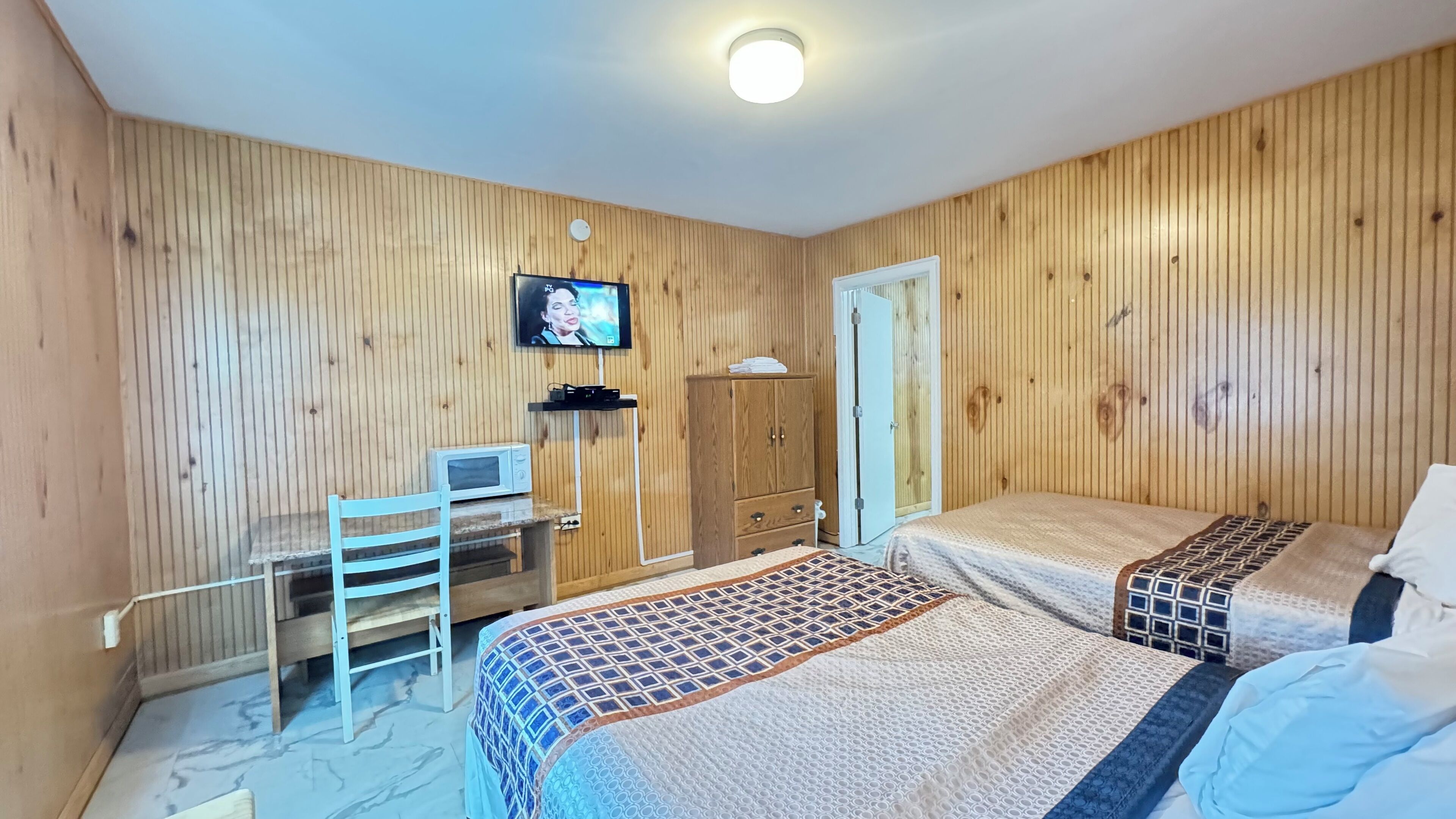 A room with wooden panel walls, a bed with patterned bedding, a desk with a computer, and a television mounted on the wall.