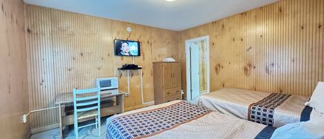 A room with wooden panel walls, a bed with patterned bedding, a desk with a computer, and a television mounted on the wall.