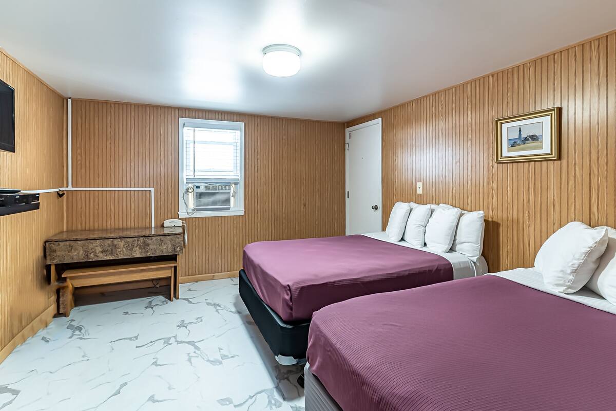 A room with wooden paneling, two beds with purple bedding, a bench, a desk, a window, and a framed picture on the wall.