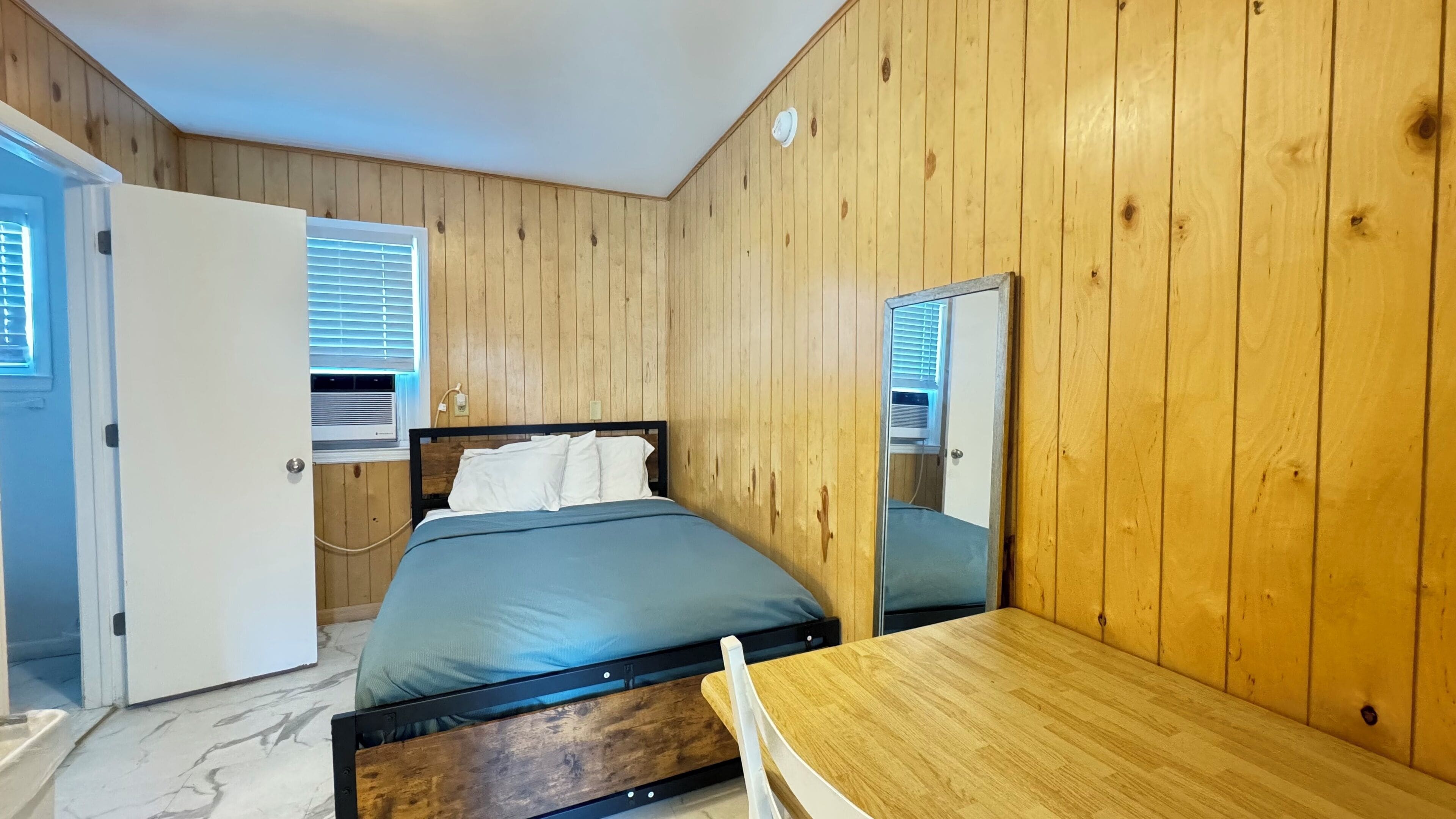 A bedroom with a wooden paneled wall, a bed with a blue mattress, a wooden table, and a mirror.