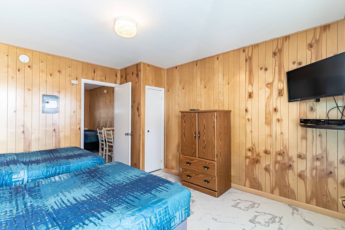 A bedroom with wooden panel walls, a bed with a blue patterned bedspread, a wooden wardrobe, and a mounted TV.