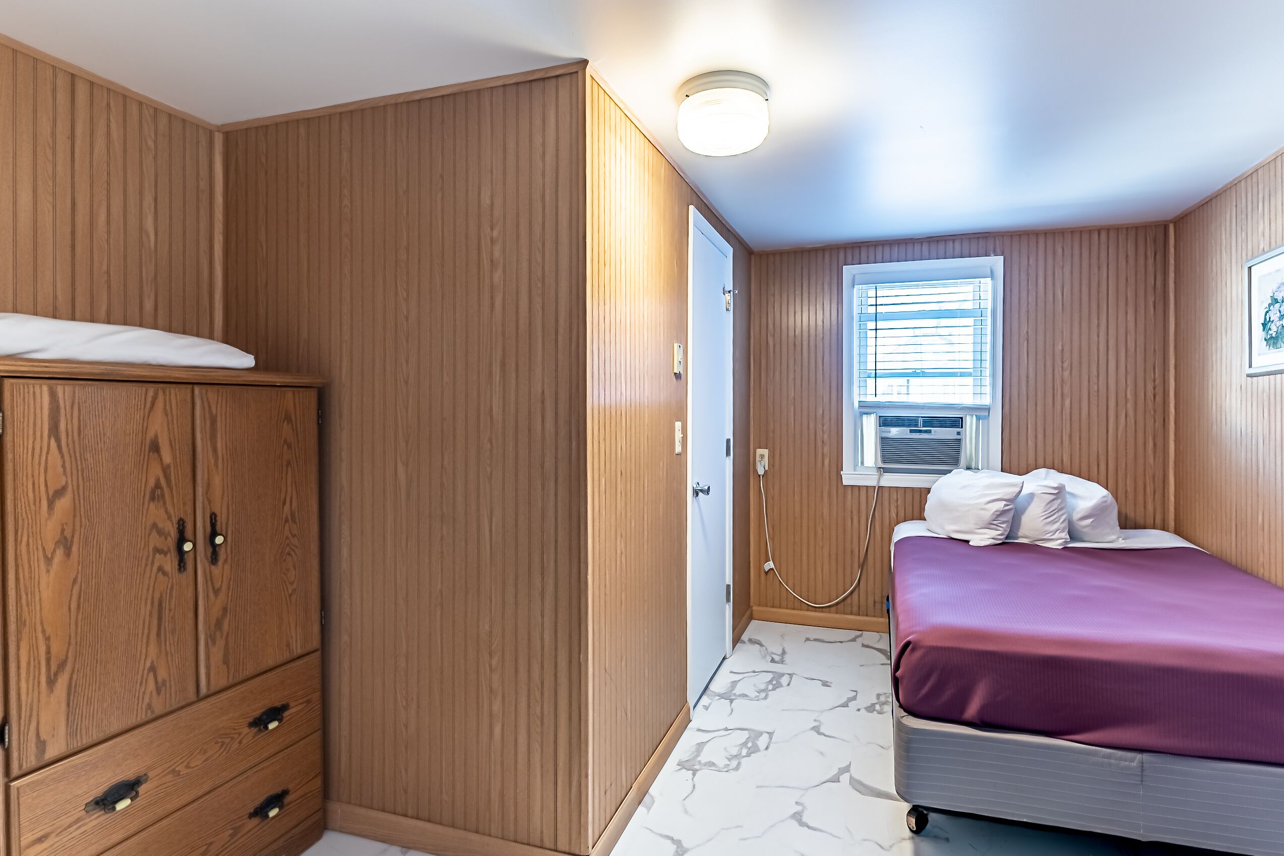 Une chambre avec des lambris en bois, un lit recouvert d’une courtepointe violette, une armoire et une fenêtre avec des stores.