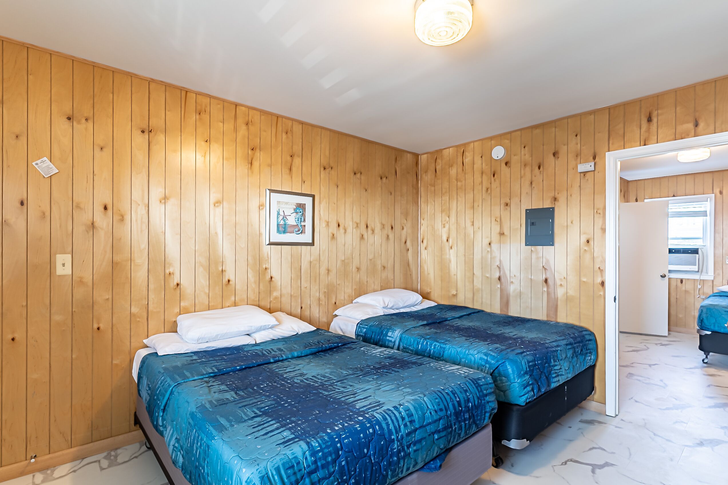 A room with wooden panel walls, two beds with blue bedding, a framed picture on the wall, and a door leading to another room.