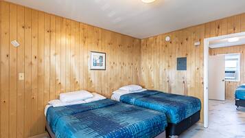 A room with wooden panel walls, two beds with blue bedding, a framed picture on the wall, and a door leading to another room.