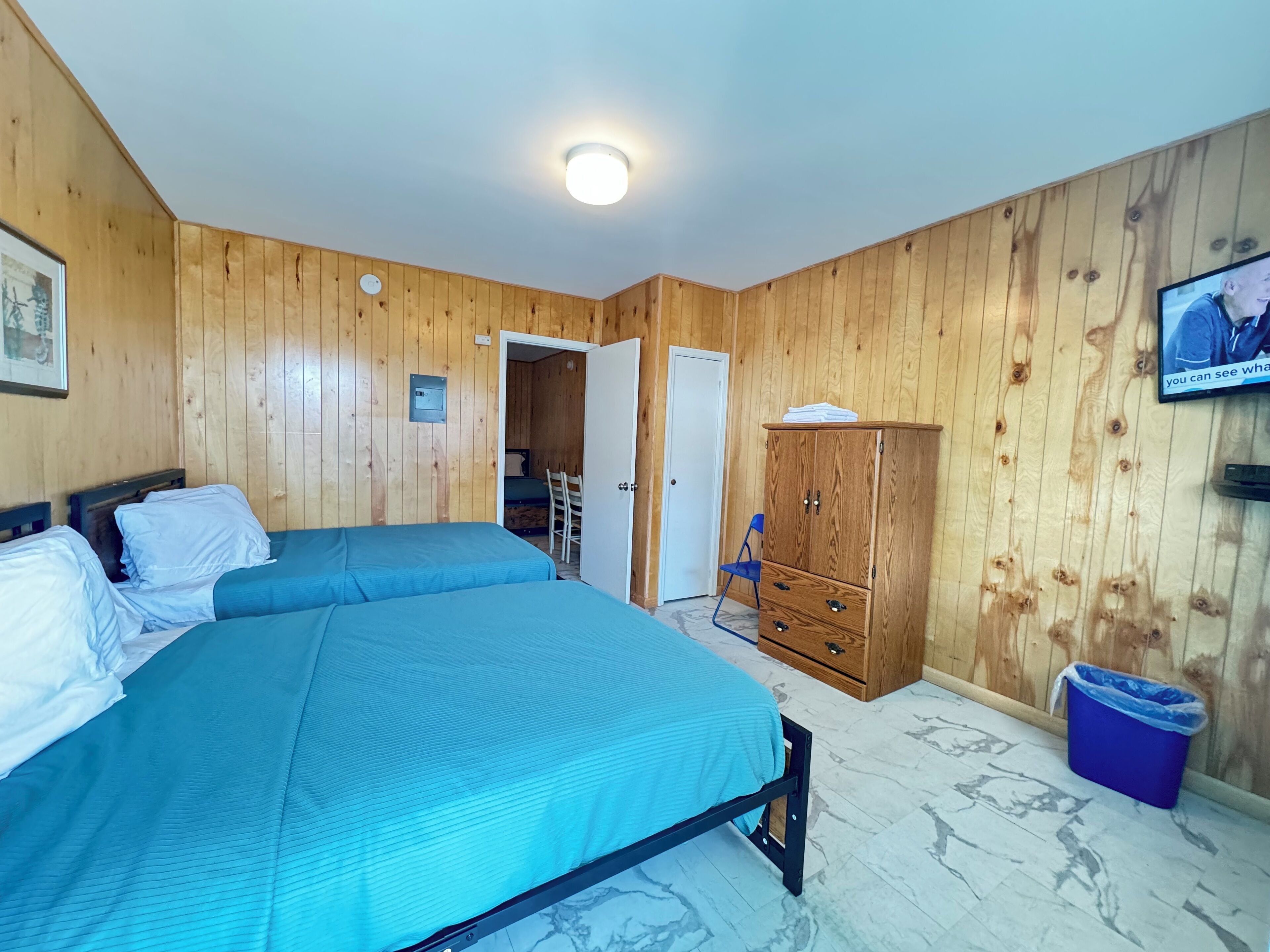 Una habitación con paneles de madera, una cama con ropa de cama azul, un armario de madera, un televisor y una puerta que da a otra habitación.