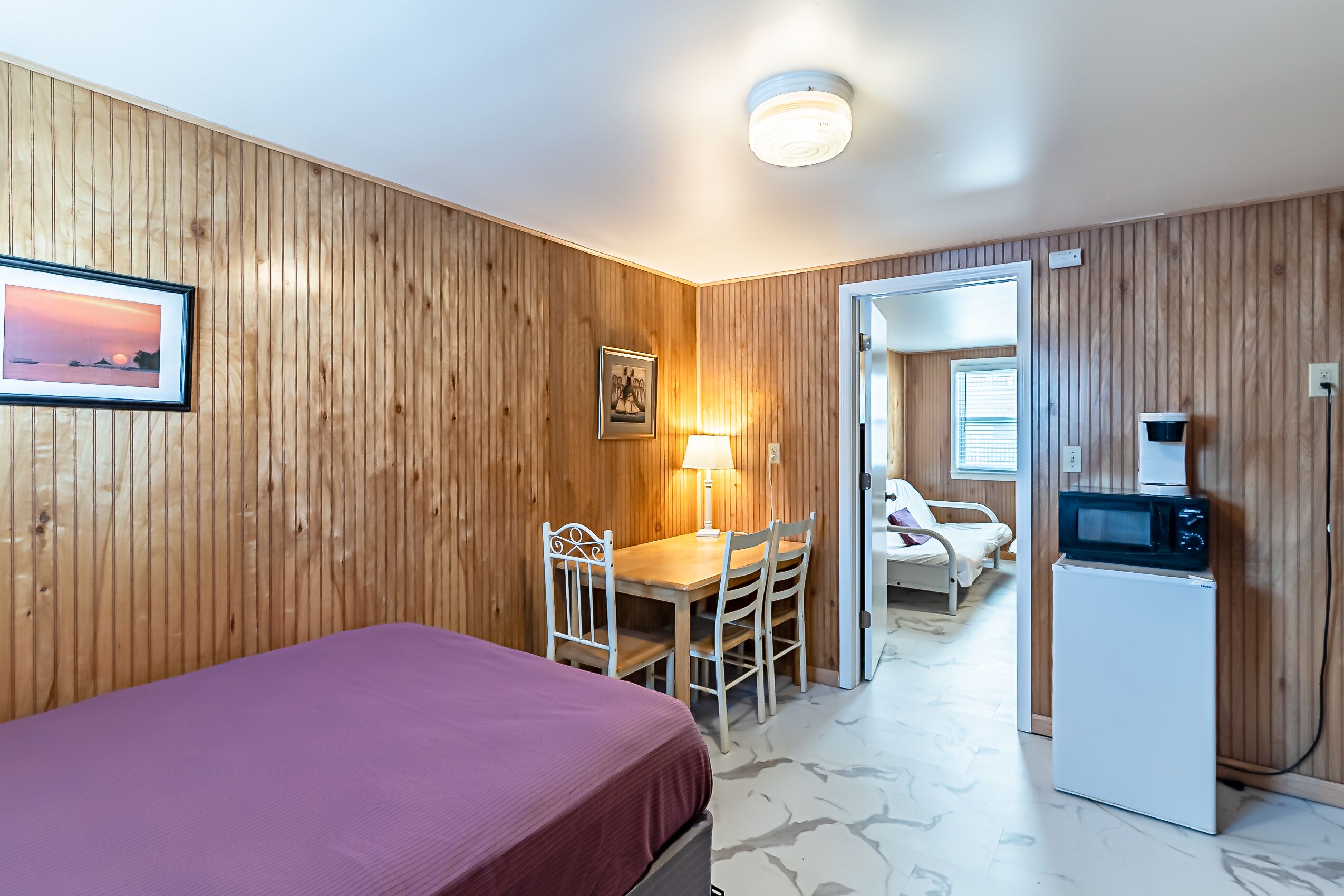 A room with wooden paneling, a bed, a desk with a chair, a lamp, a microwave, and a door leading to another room.