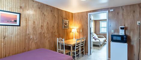 A room with wooden paneling, a bed, a desk with a chair, a lamp, a microwave, and a door leading to another room.