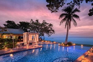 Un área de piscina lujosa con un pabellón, palmeras y vistas al océano al atardecer.
