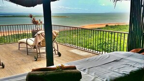 A balcony with a bed, a chair, and a cart, overlooking a beach and ocean. - Bumi Hills Safari Lodge (Bumi Hills)
