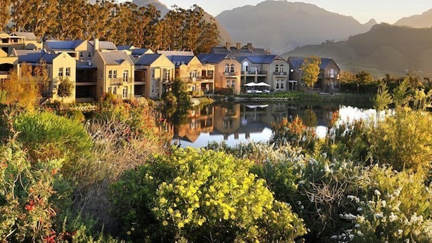 A row of traditional-style buildings with a reflective pond in the foreground, surrounded by lush vegetation and mountains in the background. - L Ermitage Franschhoek Chateau Villas (Franschhoek)