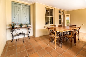 Un coin repas avec une table en bois et des chaises en osier, des plantes en pot sur une étagère et une fenêtre avec un volet bleu.