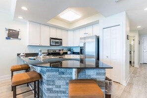 A modern kitchen with white cabinets, a built-in microwave, a stainless steel refrigerator, and a stone island with bar stools. - Beachfront Condo with Stunning Gulf Front Views | Pool & Sauna | Sleeps 8 (Gulf Shores)