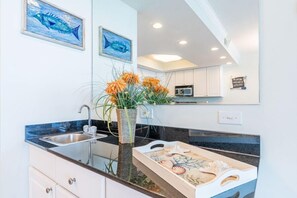 A modern kitchen with a granite countertop, stainless steel sink, and a vase with orange flowers. There are two framed paintings on the wall and a microwave in the background. - Beachfront Condo with Stunning Gulf Front Views | Pool & Sauna | Sleeps 8 (Gulf Shores)