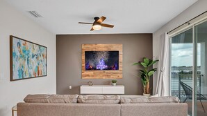 Un salon moderne avec un canapé gris, un ventilateur de plafond, un téléviseur fixé au mur diffusant des images de feux d’artifice et un balcon avec vue.