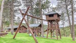 Una estructura de madera rústica con una torre de vigilancia, rodeada por un césped cuidado y otras construcciones de madera en un entorno boscoso.