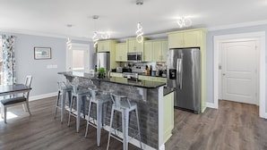 A modern kitchen with a central island, bar stools, stainless steel appliances, and light green cabinets. - Spacious Home with Private Pool, Steps from the Beach & Marsh Views (Murrells Inlet)