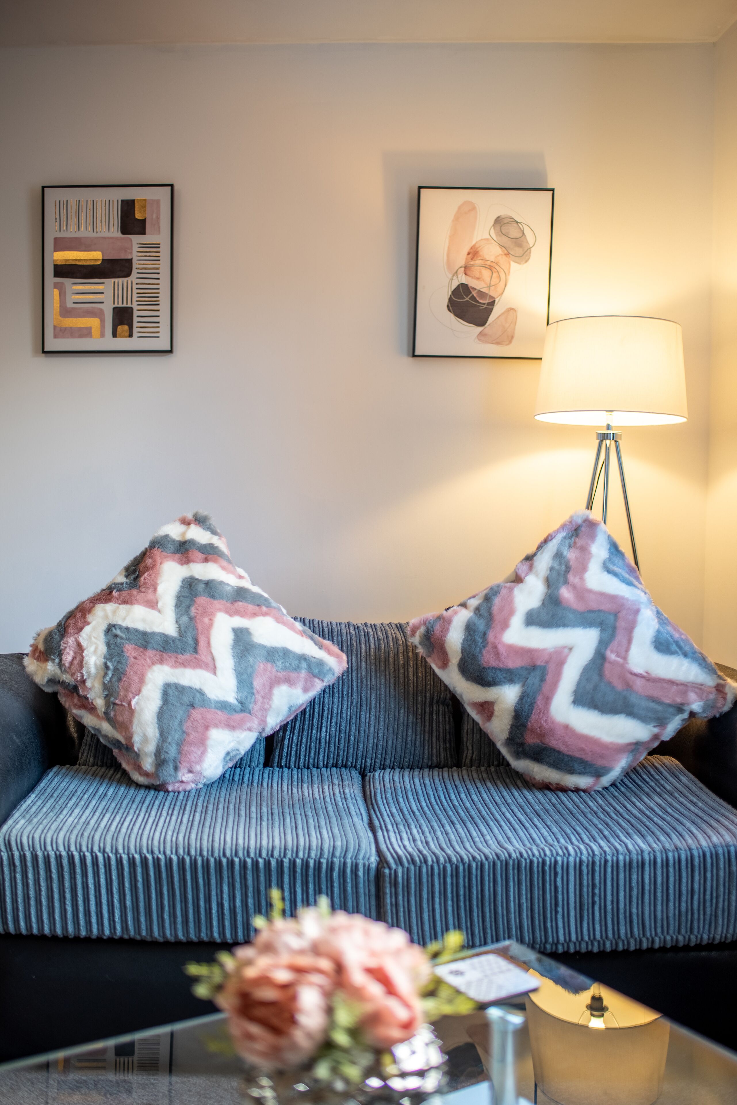 A modern living room with a striped sofa, two patterned throw pillows, a floor lamp, and a vase with flowers on a glass table.