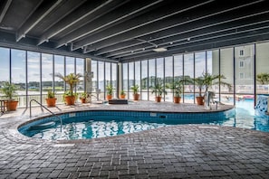 Uma piscina interior com chão de tijolos, rodeada por plantas em vasos e grandes janelas que oferecem vista para o exterior.