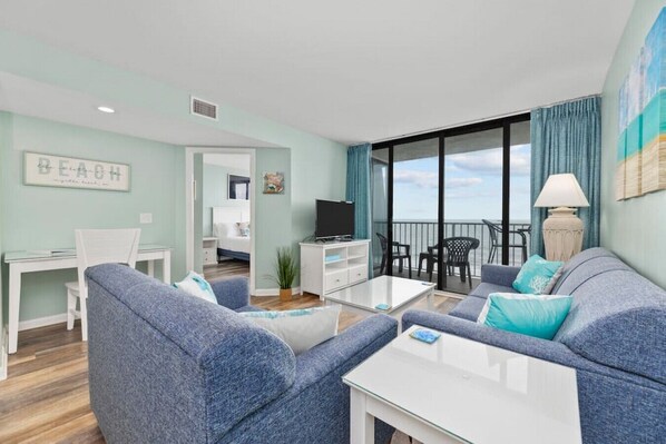 Un salon moderne avec un canapé, une table basse et un téléviseur. Il y a un balcon avec vue sur la mer.