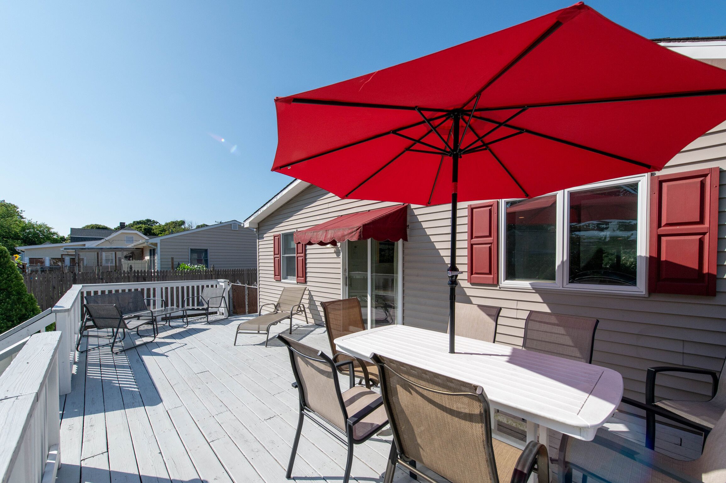 A wooden deck with a table and chairs, a red umbrella, and a house with red shutters.