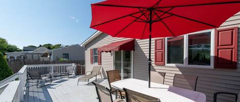 A wooden deck with a table and chairs, a red umbrella, and a house with red shutters.