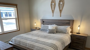 A bedroom with a wooden bed, two bedside tables with lamps, a window with a view of a building, and a wall-mounted decorative item. - Will it be ICE AXE, CRAWLER, NORDIC? 1200MGPS-Mtn Views! On River! BBQ & yard! (Ouray)